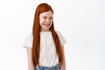 Wall Mural - Cute little redhead girl with long ginger hair and freckles, laughing and smiling, gigggle over something funny, standing against white background