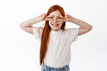 Wall Mural - Happy little girl with red long hair and freckles, smiling toothy and showing v-signs, disco gesture over eyes, having fun in playful mood, standing silly against white background