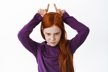 Wall Mural - Serious and determined redhead girl making bull horns gesture, holding fingers above head, look from under forehead confident and sly, white background