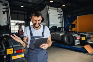 Wall Mural - Smiling tattooed bearded blue collar worker in overalls using tablet to check on delivery while standing in garage of import and export firm. In background are trucks.