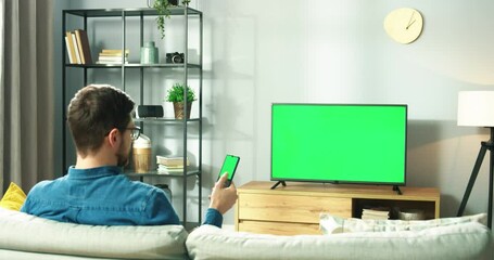 Caucasian young man in glasses sitting on sofa in room sitting in house watching TV with green screen and tapping on cellphone with chroma key searching internet using social network, back view