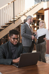 Wall Mural - African businessman in eyeglasses sitting at the table and working on laptop with business people talking in the background