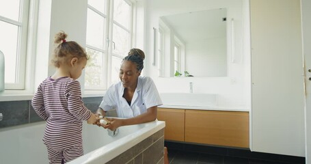 Wall Mural - Smiling young African mom having fun with her little girl in a bathtub while teaching her how to brush her teeth