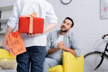 Wall Mural - back view of child holding gift box and happy fathers day card near amazed arabian dad on blurred background