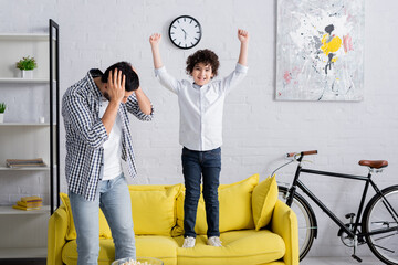 Wall Mural - excited arabian boy jumping on sofa near tired father covering head with hands