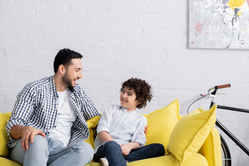 Wall Mural - happy arabian father and son talking while sitting on sofa at home