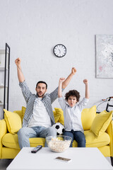 Wall Mural - excited arabian kid and father showing success gesture while watching football match at home