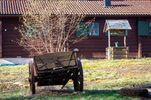 Old Traditional Swedish Courtyard With Vintage Style Well Near A Red Wooden House. Wooden Farm Cart With Big Wheels. Countryside Early Spring In Scandinavia. Traditional Swedish Farm Buildings.