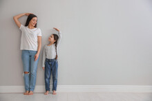 Little Girl And Mother Measuring Their Height Near Light Grey Wall Indoors. Space For Text