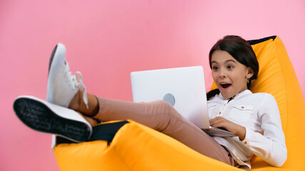 amazed child using laptop while resting on bean bag chair isolated on pink