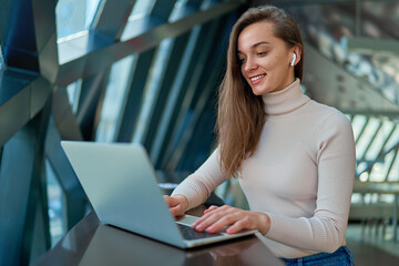 Wall Mural - Young modern casual happy joyful smart woman wearing wireless headphones using laptop for watching video and remote working online while sitting and resting at cafe