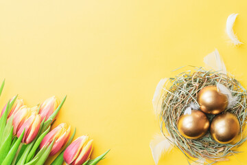 Wall Mural - Golden easter colour eggs in basket with spring tulips, white feathers on pastel yellow background in Happy Easter decoration. Spring holiday top view concept.