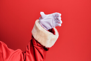 Hand of a man wearing santa claus costume and gloves over red background holding invisible object, empty hand doing clipping and grabbing gesture