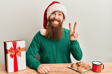 Sticker - Young irish redhead man wearing christmas sitting on the table with gift smiling with happy face winking at the camera doing victory sign. number two.