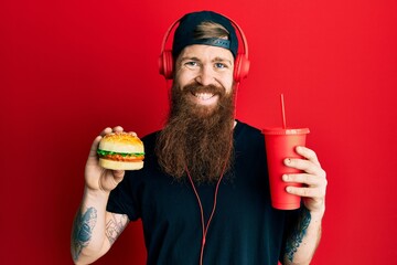 Sticker - Redhead man with long beard eating a tasty classic burger and drinking soda smiling with a happy and cool smile on face. showing teeth.