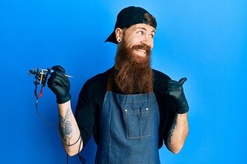 Canvas Print - Redhead man with long beard tattoo artist wearing professional uniform and gloves smiling with happy face looking and pointing to the side with thumb up.