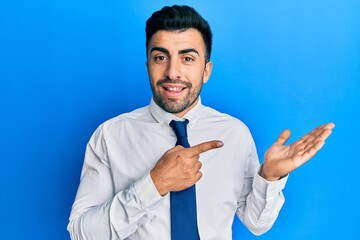 Wall Mural - Young hispanic man wearing business clothes amazed and smiling to the camera while presenting with hand and pointing with finger.