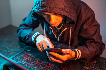 Wall Mural - Hacker with phone is typing on a laptop keyboard in a dark room under a neon light. Cybercrime fraud and identity theft