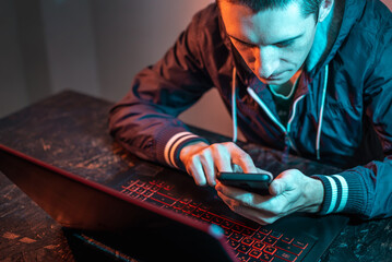 Wall Mural - Hacker with phone is typing on a laptop keyboard in a dark room under a neon light. Cybercrime fraud and identity theft