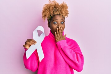 Canvas Print - Young african woman with afro hair holding pink cancer ribbon covering mouth with hand, shocked and afraid for mistake. surprised expression