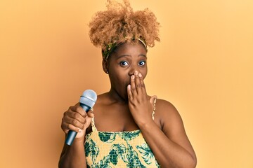 Canvas Print - Young african woman with afro hair singing song using microphone covering mouth with hand, shocked and afraid for mistake. surprised expression