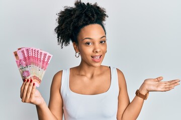 Poster - Young african american girl holding indonesian rupiah banknotes celebrating achievement with happy smile and winner expression with raised hand