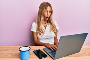 Canvas Print - Beautiful blonde young woman working using computer laptop scared and amazed with open mouth for surprise, disbelief face