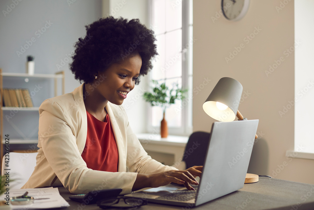 Young afro american businesswoman working on laptop with electronic documents sitting in the office. Smiling woman typing on the keyboard by sending an email or filling out an electronic database. - obrazy, fototapety, plakaty 