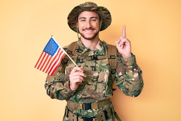 Wall Mural - Young caucasian man wearing camouflage army uniform holding usa flag surprised with an idea or question pointing finger with happy face, number one