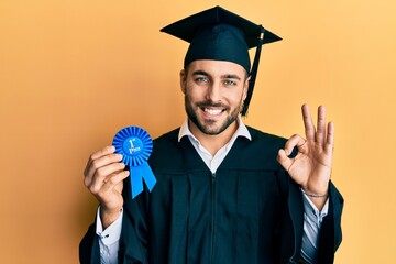 Young hispanic man wearing graduation robe holding 1 st place badge doing ok sign with fingers, smiling friendly gesturing excellent symbol