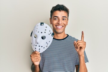 Wall Mural - Young handsome african american man holding hockey mask smiling with an idea or question pointing finger with happy face, number one