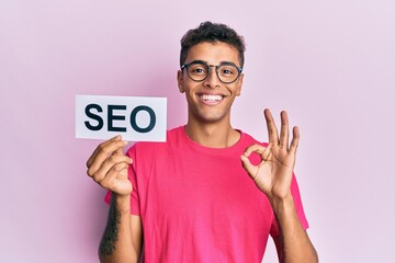 Canvas Print - Young handsome african american man holding seo message paper doing ok sign with fingers, smiling friendly gesturing excellent symbol