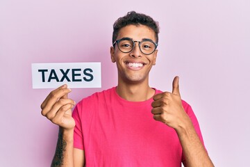 Sticker - Young handsome african american man holding taxes word smiling happy and positive, thumb up doing excellent and approval sign