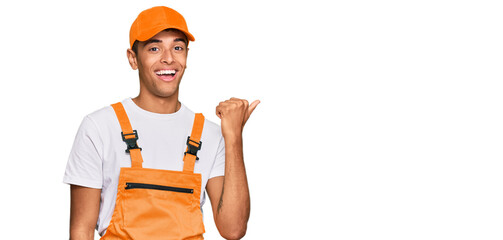 Young handsome african american man wearing handyman uniform smiling with happy face looking and pointing to the side with thumb up.