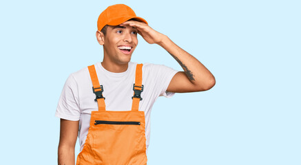 Wall Mural - Young handsome african american man wearing handyman uniform very happy and smiling looking far away with hand over head. searching concept.