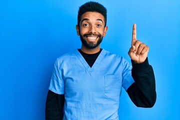 Canvas Print - Handsome hispanic man with beard wearing blue male nurse uniform pointing finger up with successful idea. exited and happy. number one.