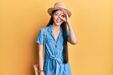 Canvas Print - Young chinese woman wearing summer hat doing ok gesture with hand smiling, eye looking through fingers with happy face.