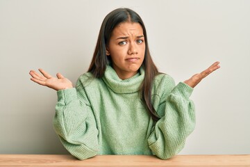 Sticker - Beautiful hispanic woman wearing casual clothes sitting on the table clueless and confused with open arms, no idea and doubtful face.