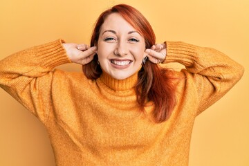 Wall Mural - Beautiful redhead woman wearing casual winter sweater over yellow background smiling pulling ears with fingers, funny gesture. audition problem