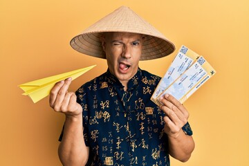 Wall Mural - Middle age bald man wearing traditional asian hat holding boarding pass winking looking at the camera with sexy expression, cheerful and happy face.