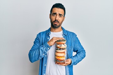 Sticker - Young hispanic man holding tasty colorful doughnuts in shock face, looking skeptical and sarcastic, surprised with open mouth