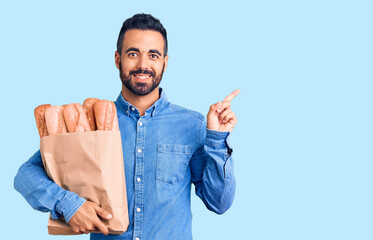 Wall Mural - Young hispanic man holding bag with bread smiling happy pointing with hand and finger to the side