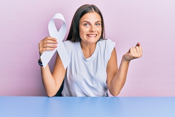 Young caucasian woman holding white ribbon screaming proud, celebrating victory and success very excited with raised arm