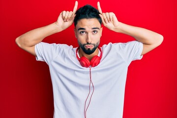 Young man with beard listening to music using headphones doing funny gesture with finger over head as bull horns