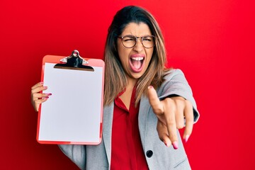 Canvas Print - Beautiful brunette woman holding clipboard with blank space angry and mad screaming frustrated and furious, shouting with anger. rage and aggressive concept.