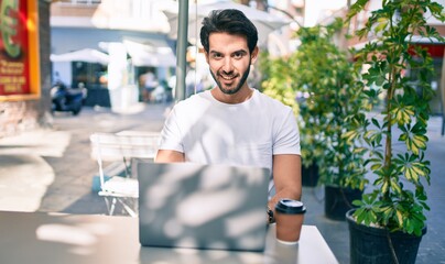 Wall Mural - Young hispanic man smiling happy working using laptop at coffee shop terrace.