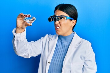 Poster - Beautiful brunette jeweller woman holding brilliant diamond stone wearing magnifier glasses clueless and confused expression. doubt concept.