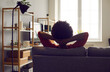 © Studio Romantic - Spending peaceful weekend on comfortable couch at home. Happy relaxed African American woman sitting hands behind head on sofa feeling safe, looking out window, contemplating future, backlit back view