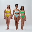 Full length shot of three attractive diverse women in colorful underwear holding hands, looking at camera, standing together isolated over light background