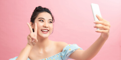 Wall Mural - Image of beautiful brunette woman laughing and showing peace sign while taking selfie photo on cellphone isolated over pink background